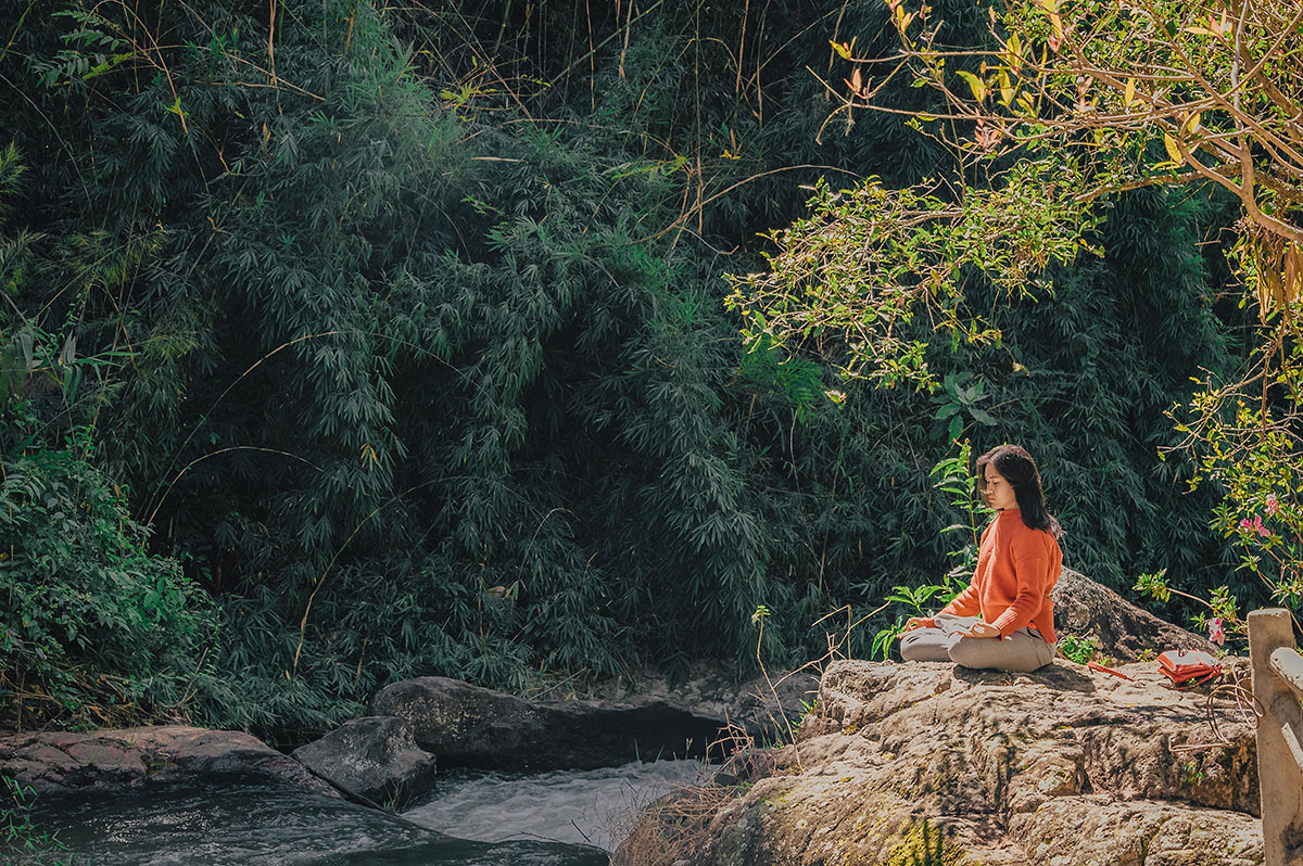 Woman in an orange shirt meditating on a rock in the forest